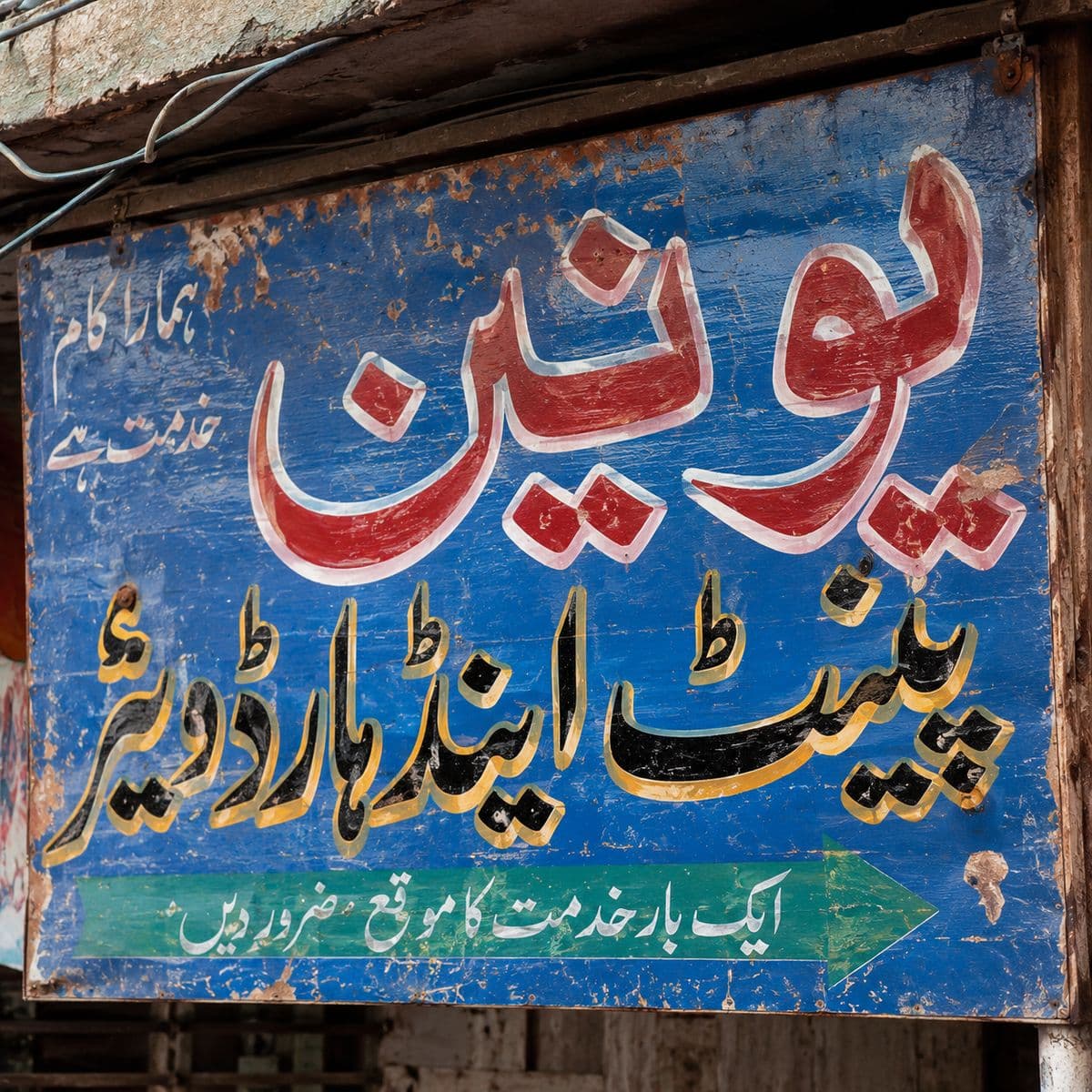 A weathered enamel signboard hand-lettered in Urdu for a paint and hardware shop.