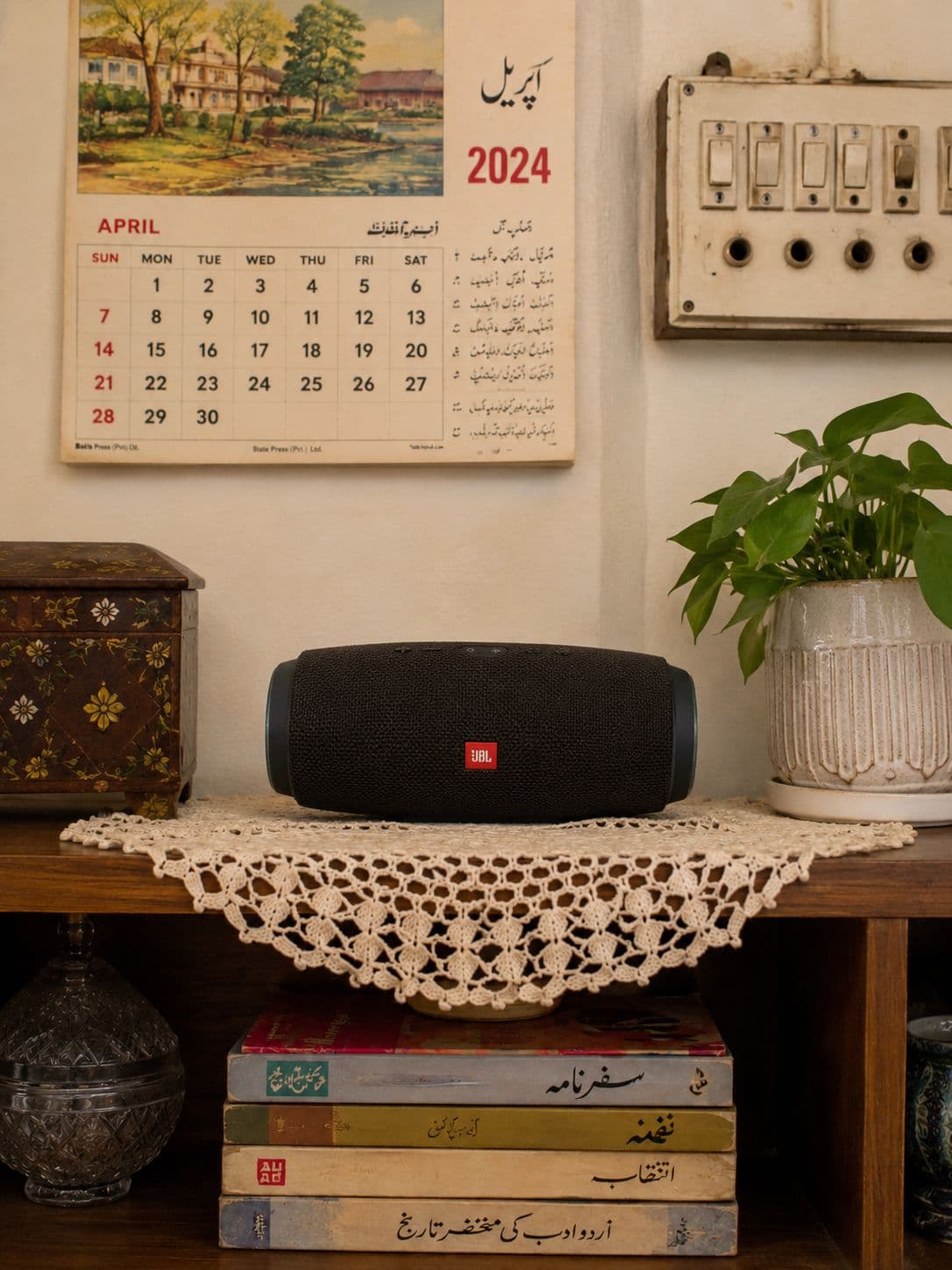 A portable Bluetooth speaker on the same crocheted doily and stack of books, beside a wall calendar open at April.